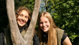 Zwei junge Frauen im Wald, die an Baumstamm stehen und freundlich in die Kamera schauen  