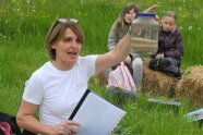 Kinder sitzen im Halbkreis auf Strohballen auf einer Wiese, eine Landwirtin erklärt etwas