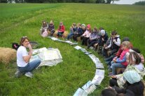 Kinder sitzen im Halbkreis auf Strohballen auf einer Wiese, eine Landwirtin erklärt etwas
